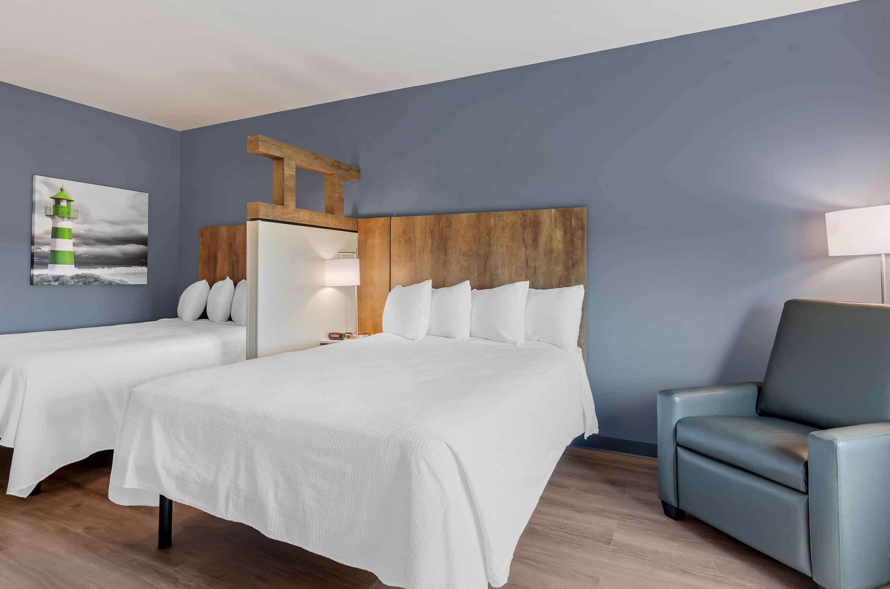 Une chambre d’hôtel avec deux lits, une tête de lit en bois, un fauteuil et un tableau représentant un phare.