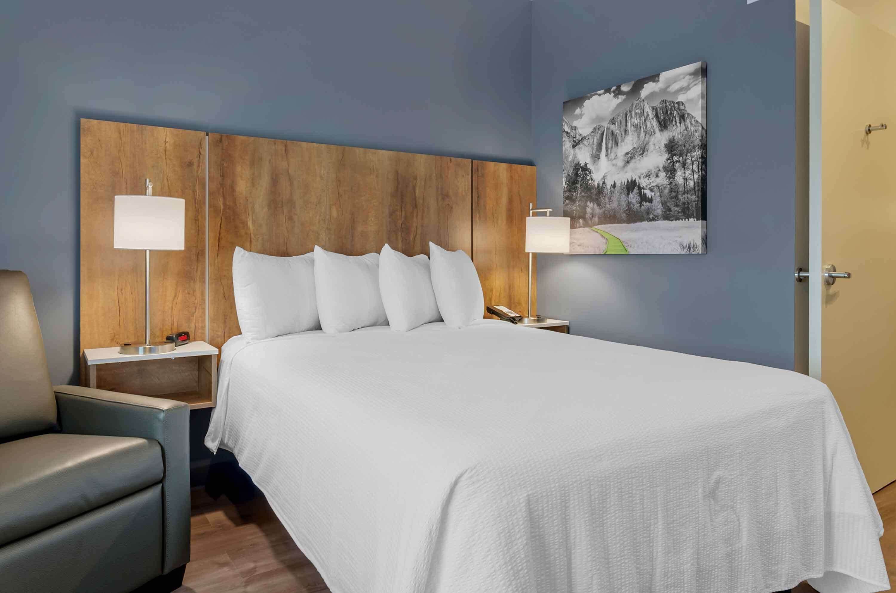 A hotel room with a large bed, two bedside tables, a chair, a painting on the wall, and a door.