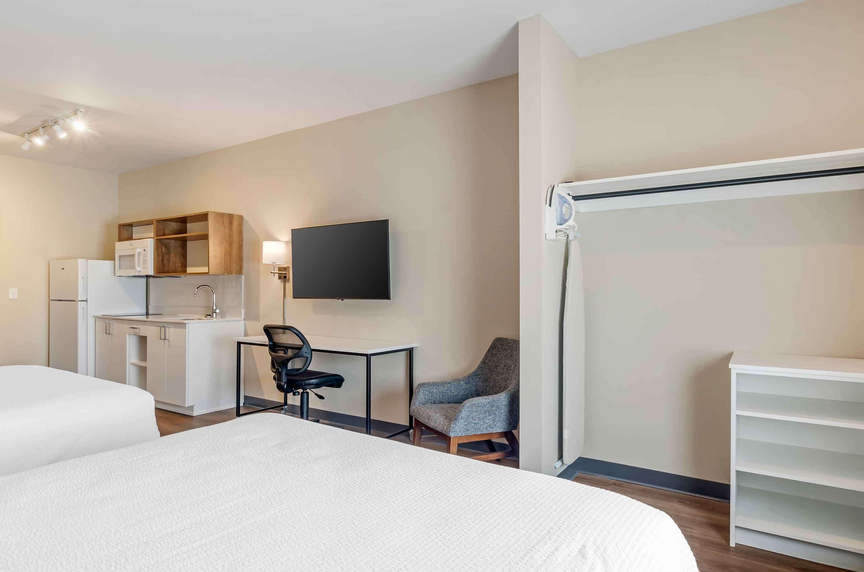 A hotel room with a bed, a desk with a chair, a television, a kitchenette, and a wardrobe.
