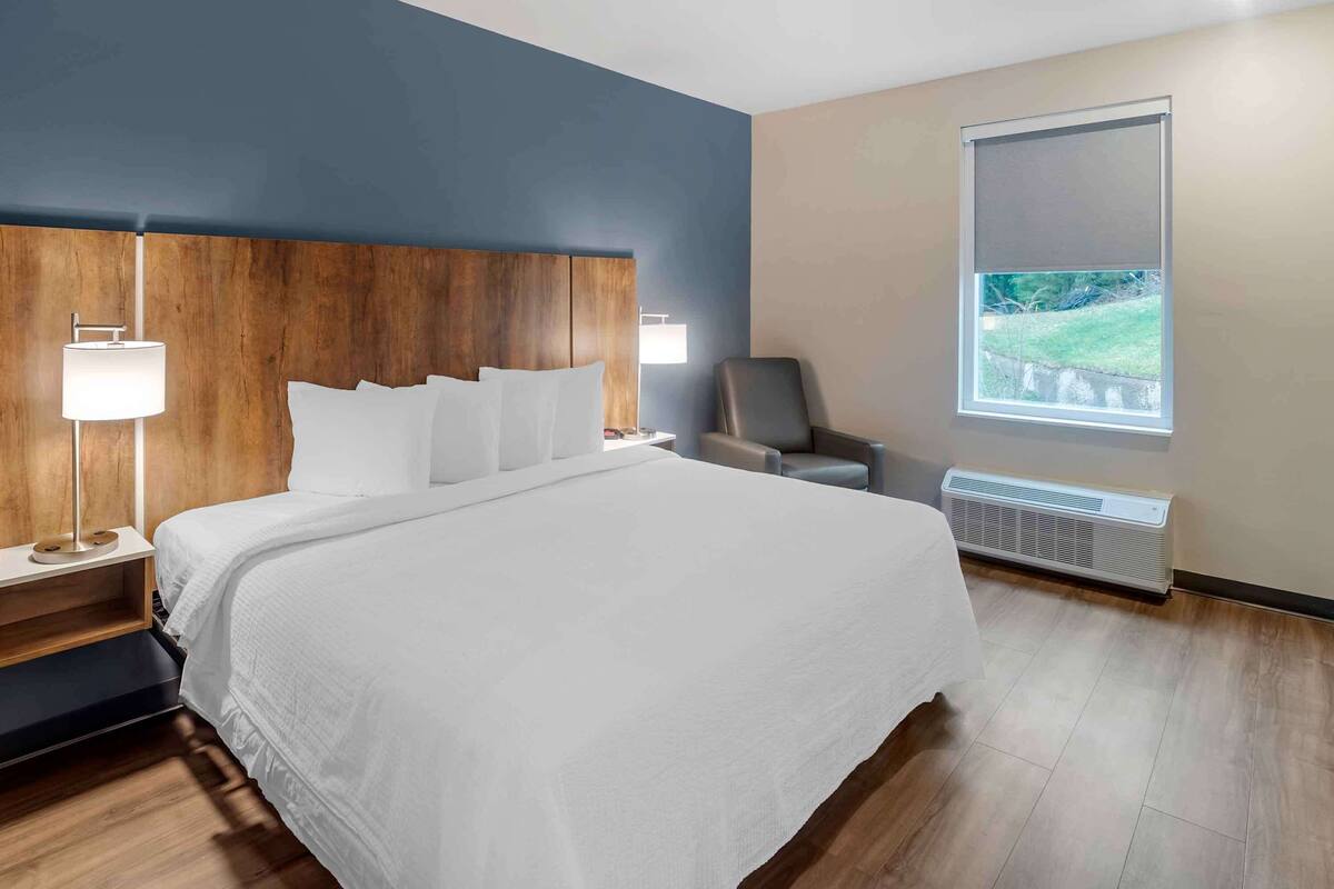 Habitación de hotel moderna con una cama grande, cabecera de madera, una silla, un escritorio, una lámpara y una ventana con vista a zonas verdes.