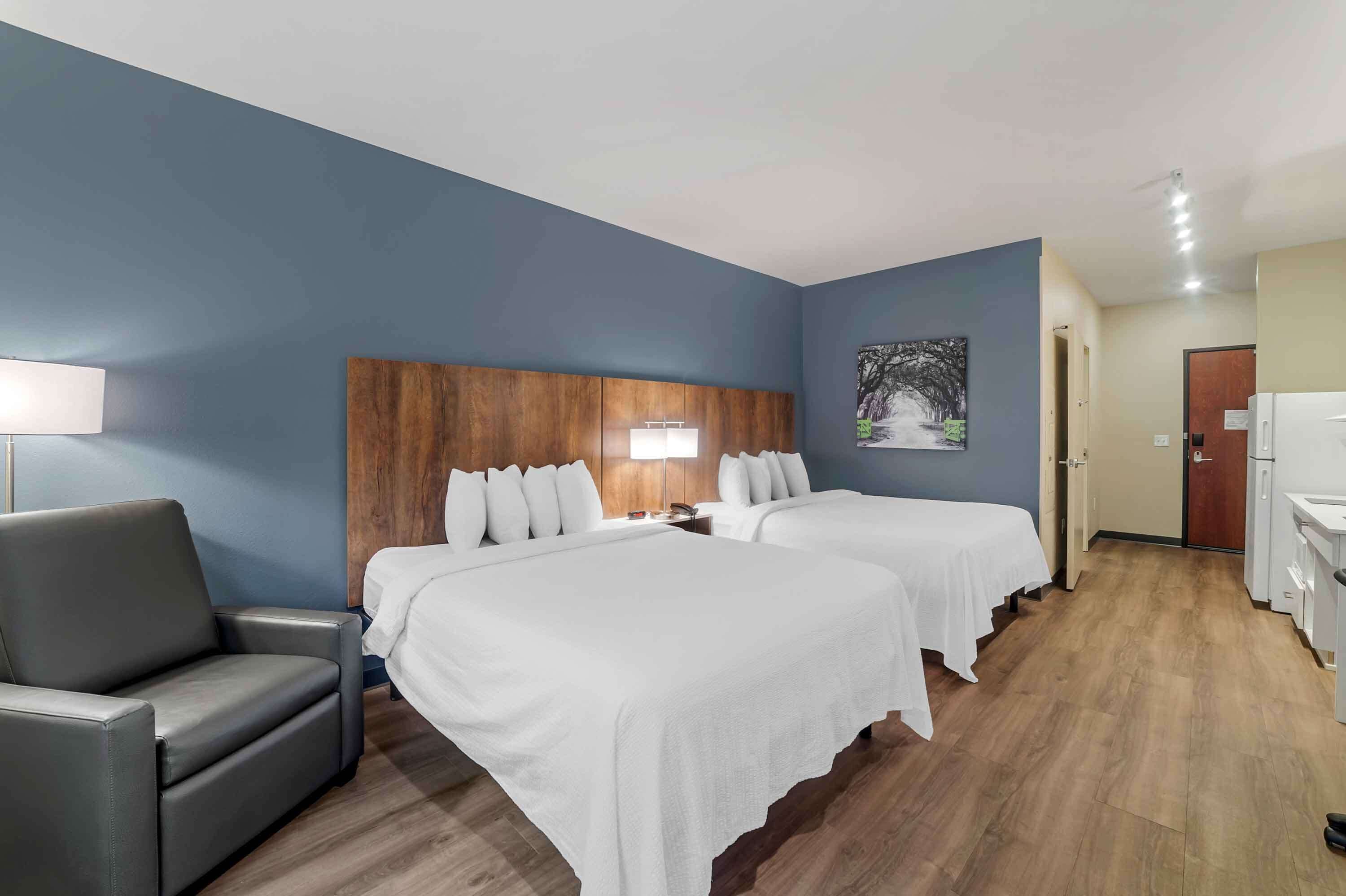 Ein Hotelzimmer mit zwei Betten, einem Holzkopfbrett, einem grauen Sessel und einem Bild an der Wand.