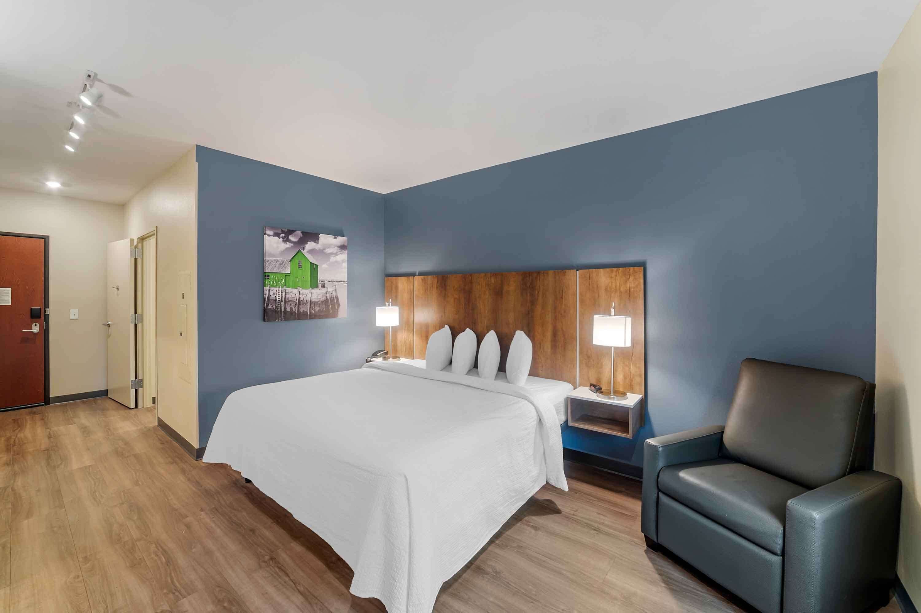 A hotel room with a bed, a chair, a nightstand, and a wall-mounted headboard.