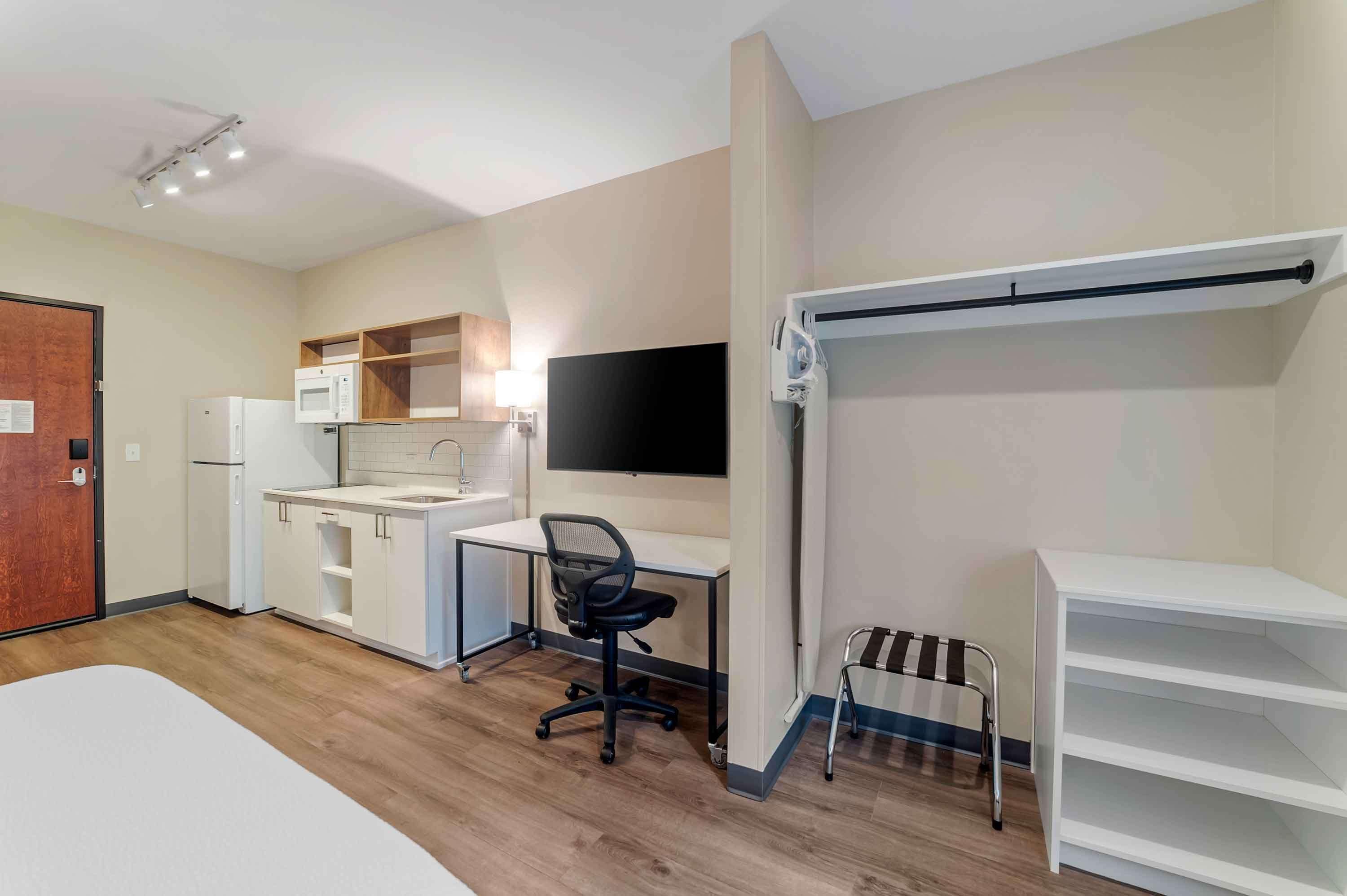 A compact room with a kitchenette, a desk with a chair, a bed, and a TV mounted on the wall.