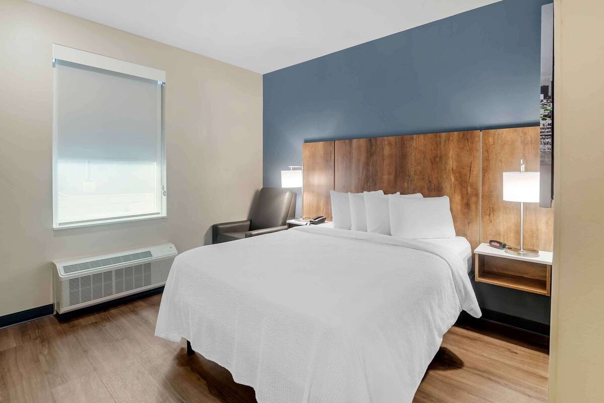 Habitación de hotel con una cama grande, una silla, una mesita de noche con lámpara y una ventana con persianas.