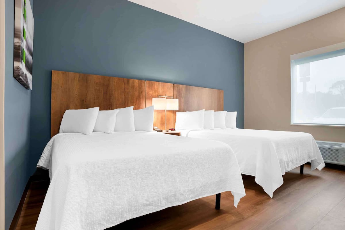 A hotel room with two beds, a wooden headboard, a nightstand with a lamp, and a large window.