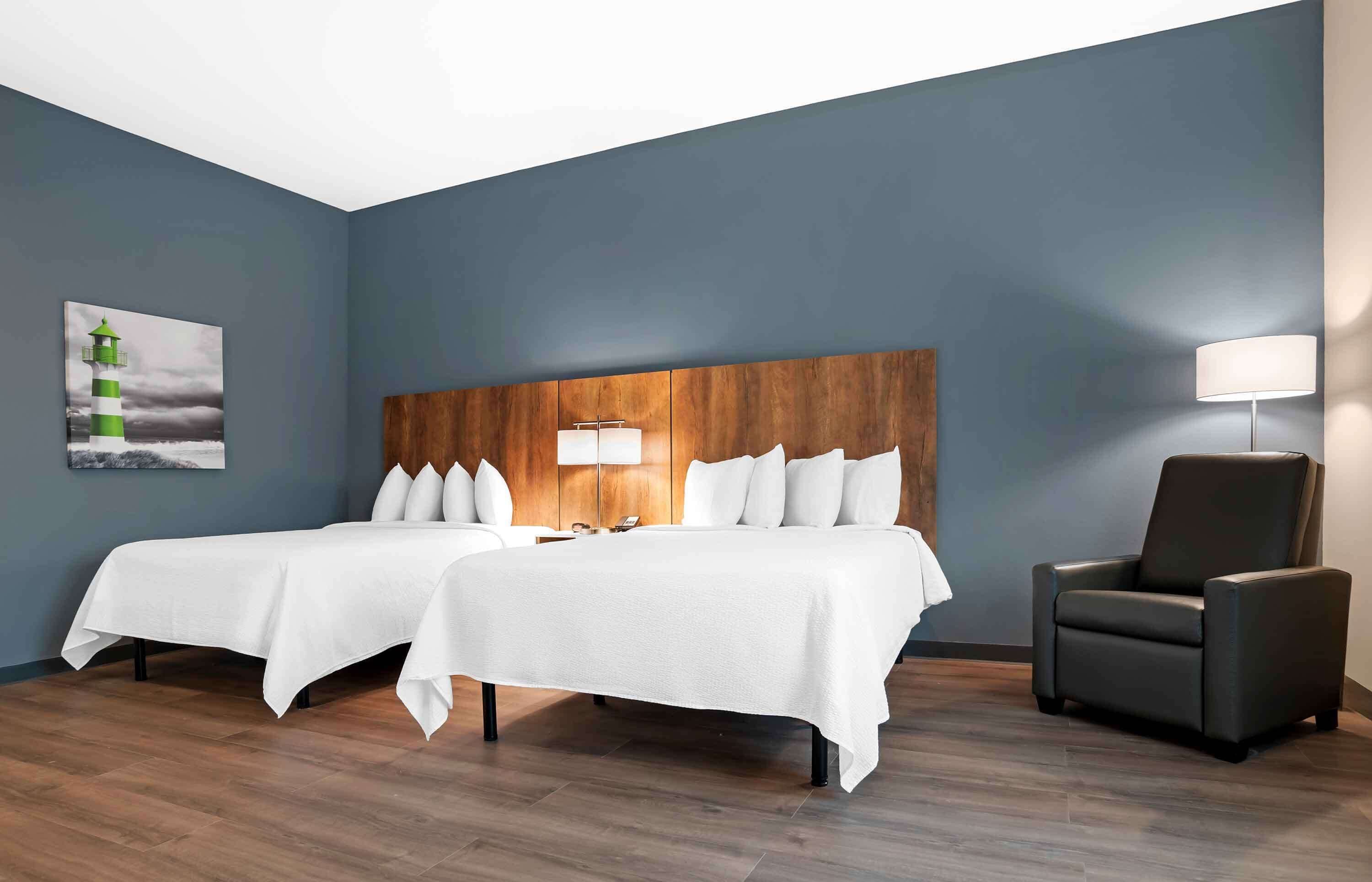 Une chambre d’hôtel avec deux lits, une tête de lit en bois, une table de chevet avec une lampe, une chaise et un tableau représentant un phare.