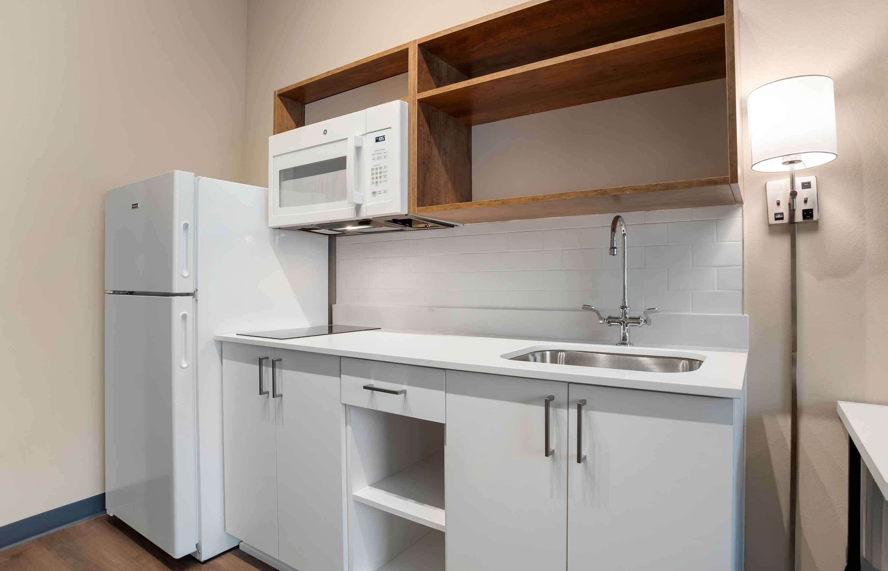 Una cocina compacta con muebles blancos, un microondas integrado, un lavamanos de acero inoxidable y un estante de madera sobre la encimera.
