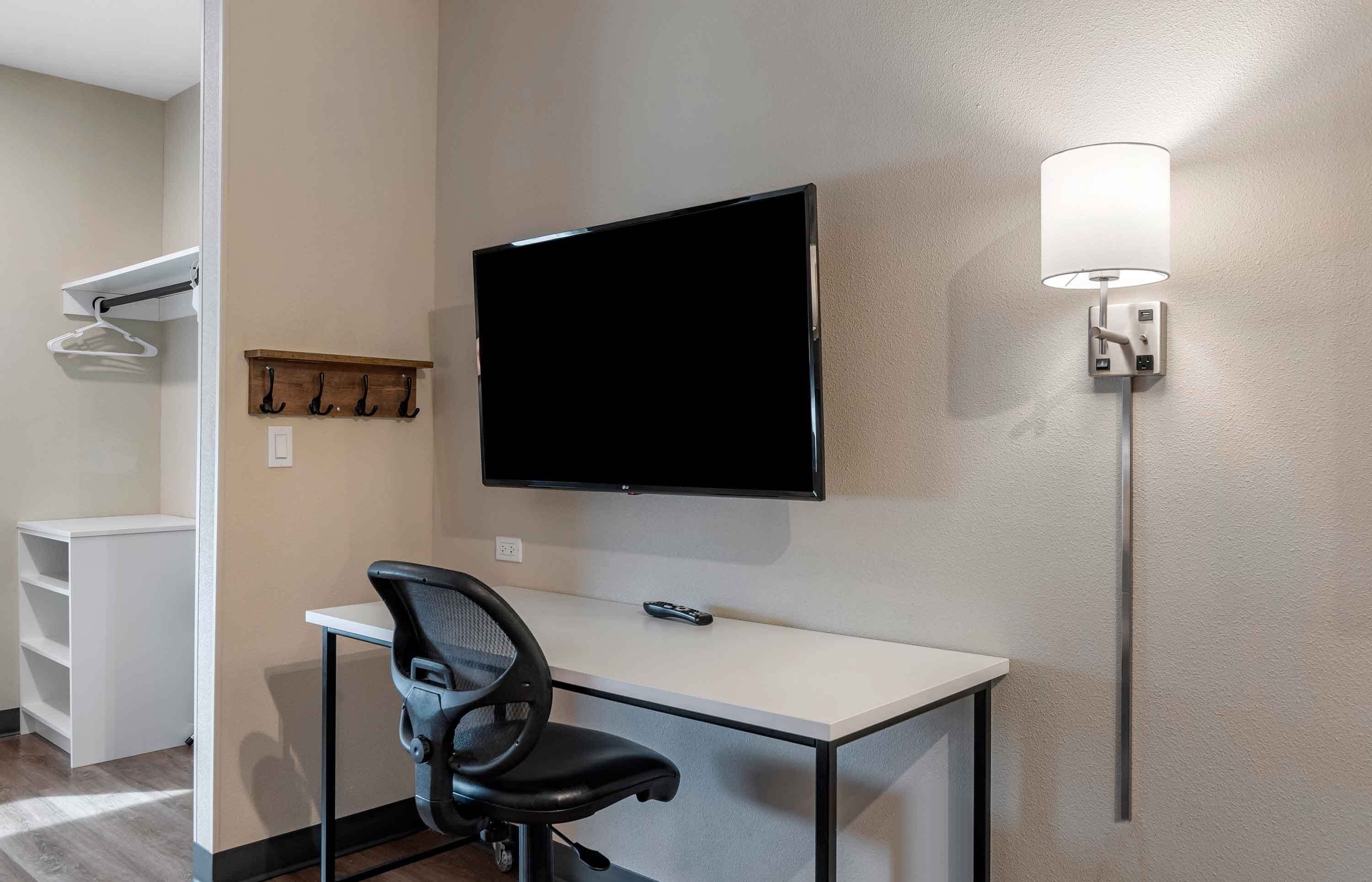 Hotelkamer met flatscreen televisie aan de muur, een bureau met stoel, een kapstok en wandlamp.