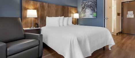 Una habitación de hotel con una cama grande, dos lámparas de noche, un sillón gris y un cabecero de madera.