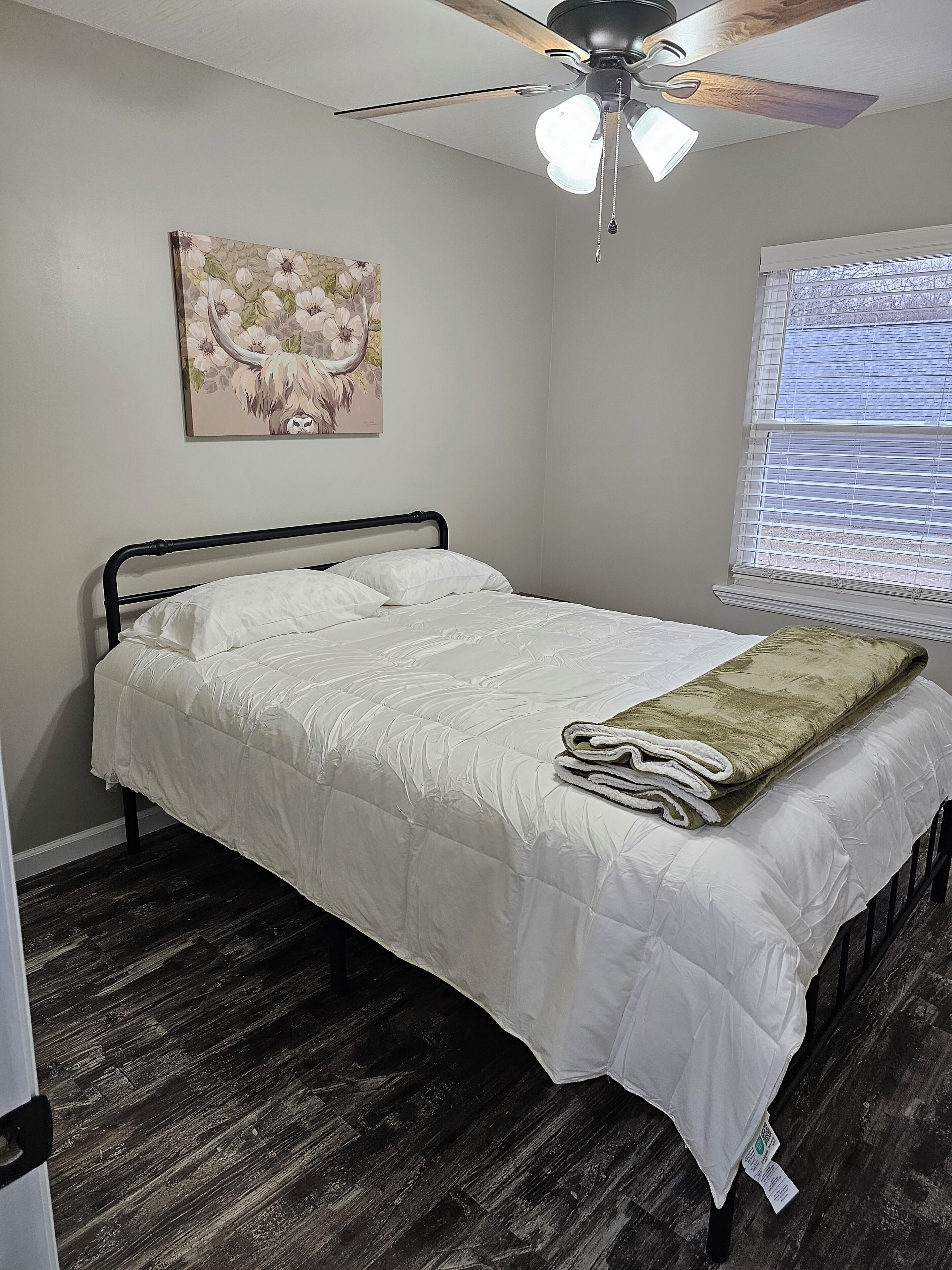 Une chambre à coucher avec un lit, un ventilateur de plafond, une fenêtre avec des stores et un tableau floral accroché au mur.