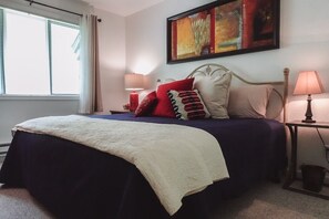 A neatly made bed with a white throw blanket, a blue bedspread, and multiple pillows in red, white, and beige. There is a painting on the wall above the bed and two bedside lamps on either side.