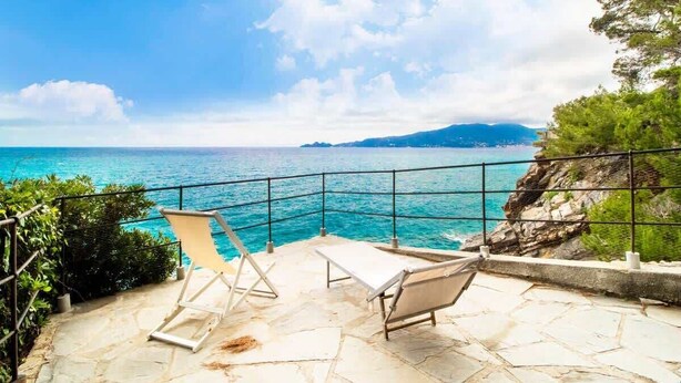A stone patio with two lounge chairs overlooking a turquoise sea and rocky coastline.