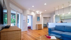 A modern living room with a blue sofa, a glass coffee table, and a wicker ottoman. There is a kitchen area with green cabinets and pendant lighting.