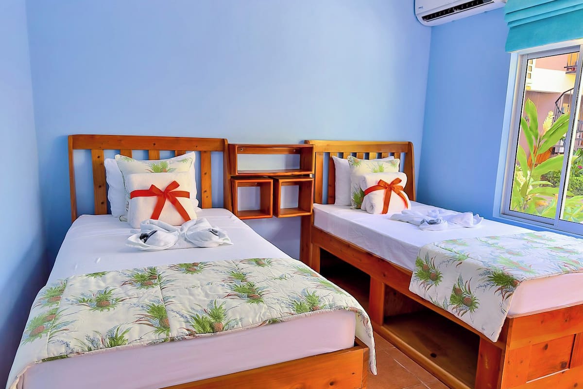 A room with two wooden beds, each with a floral bedspread and pillows tied with red ribbons. There is a small wooden table between the beds, and a window with a view of greenery outside.