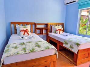 A room with two wooden beds, each with a floral bedspread and pillows tied with red ribbons. There is a small wooden table between the beds, and a window with a view of greenery outside.