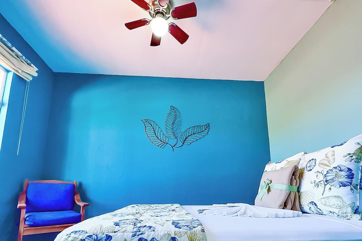 A bedroom with a bed, a chair, a ceiling fan, and a wall with a leaf design.