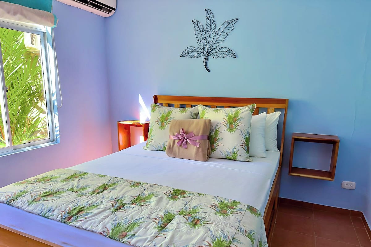 A bedroom with a bed, a nightstand, a wall decoration, and a window with a view of greenery.