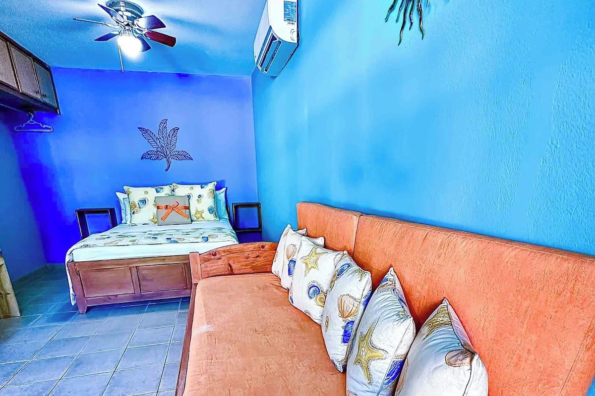 A room with a wooden sofa, a bed with patterned bedding, a ceiling fan, and a wall-mounted air conditioner.