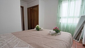 A bedroom with a patterned bedspread, a wooden wardrobe, a green curtain, and a whiteboard leaning against the wall. - 063 - Club Náutico 006 - comfortHOLIDAYS (Santa Pola)