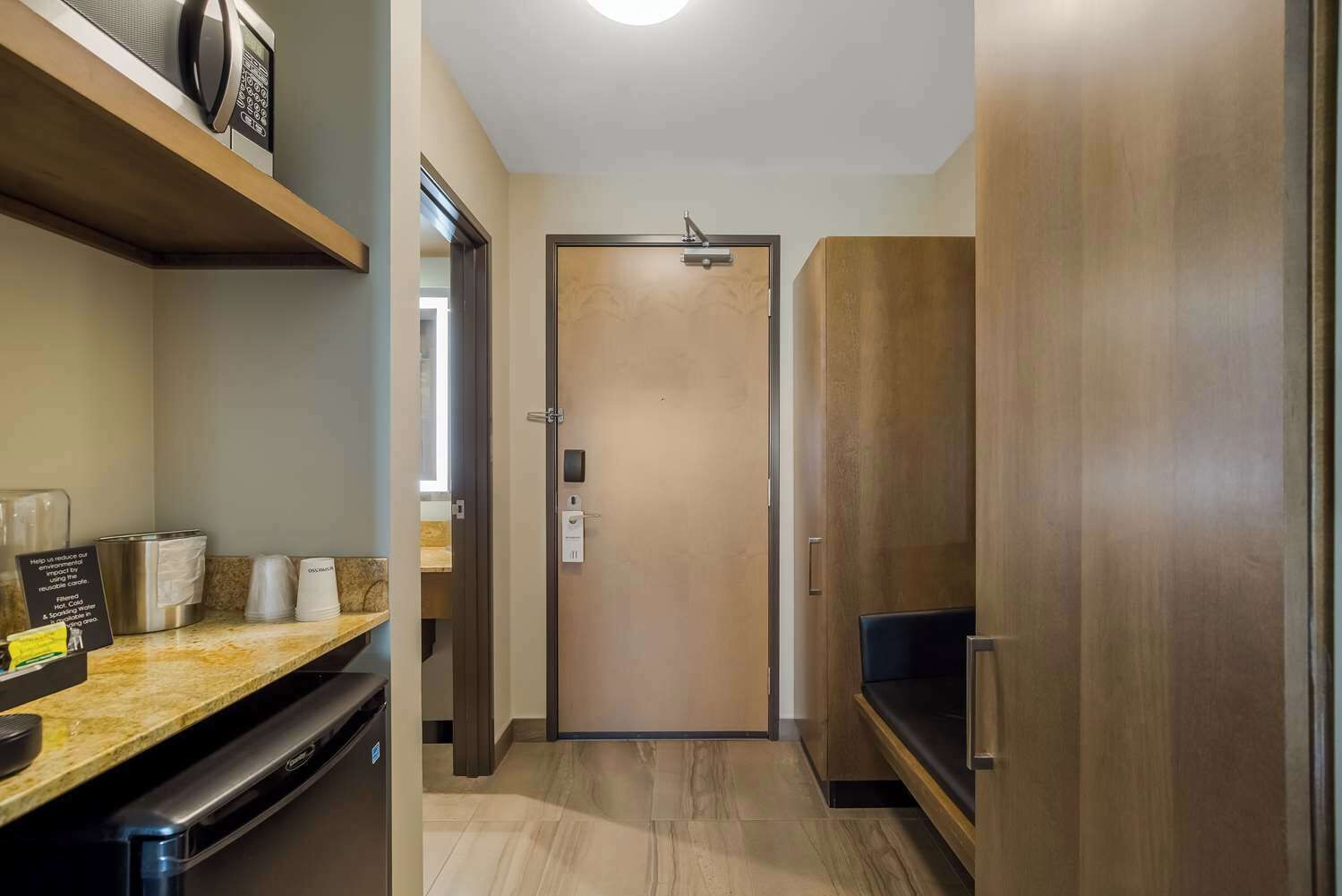 A hotel room with a wooden door, a minibar, a built-in TV, and a small seating area.