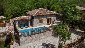 Una villa de piedra con techo de tejas, una piscina y vistas a árboles verdes exuberantes y montañas al fondo.