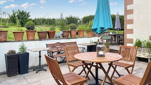 Eine Terrassengruppe im Freien mit einem runden Holztisch und Stühlen, umgeben von Topfpflanzen und einem blauen Sonnenschirm.