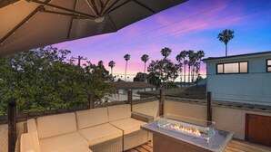 An outdoor seating area with a glass-top fire feature, surrounded by wooden decking and overlooking a residential neighborhood with palm trees and a building with a balcony.