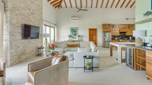A spacious living area with a stone wall, a large sofa, a coffee table, and a kitchen with wooden cabinets and a built-in refrigerator. - Marazul Belize (Placencia)