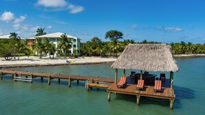 A wooden dock with a thatched-roof structure, lounge chairs, and a building with a green roof in the background. - Marazul Belize (Placencia)