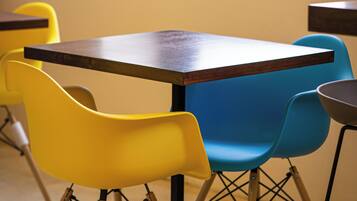 A table with a wooden top and four chairs, two yellow and two blue, in a room with a window and a plain wall.