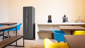 A modern kitchen with a refrigerator, a coffee machine, and a sink. There are wooden tables and chairs with blue and yellow cushions.