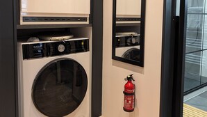Un cuarto de lavandería con una lavadora de carga frontal, un extintor de incendios sujeto a la pared y un espejo grande sobre la lavadora.