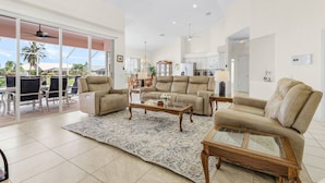 A spacious living room with a large glass door leading to a balcony, a dining area, and a kitchen in the background.