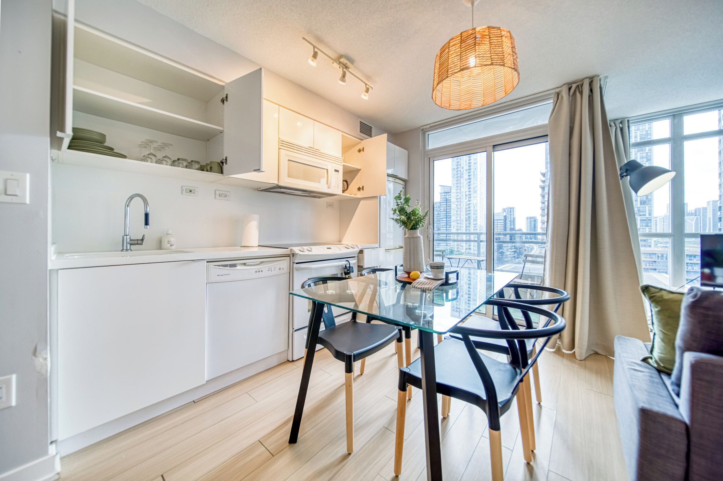 A modern kitchen with white cabinets, a glass dining table, and a city view through large windows.