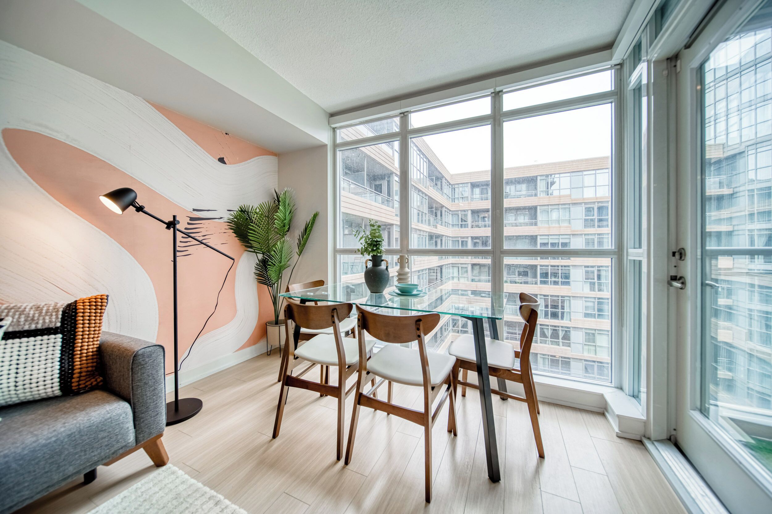 A modern living room with a glass dining table, wooden chairs, a floor lamp, and a sofa with patterned cushions.