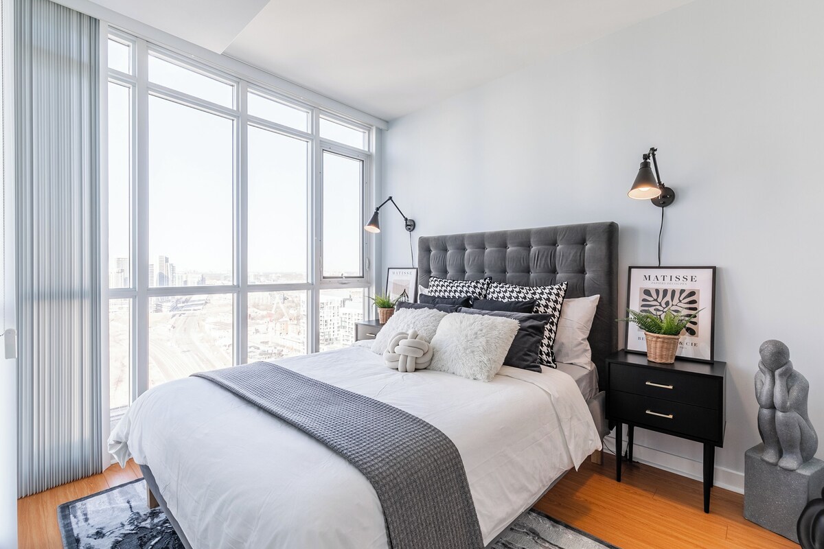 Um quarto moderno com uma cama grande, uma mesa de cabeceira, uma luminária na parede e vista para a cidade através de amplas janelas.