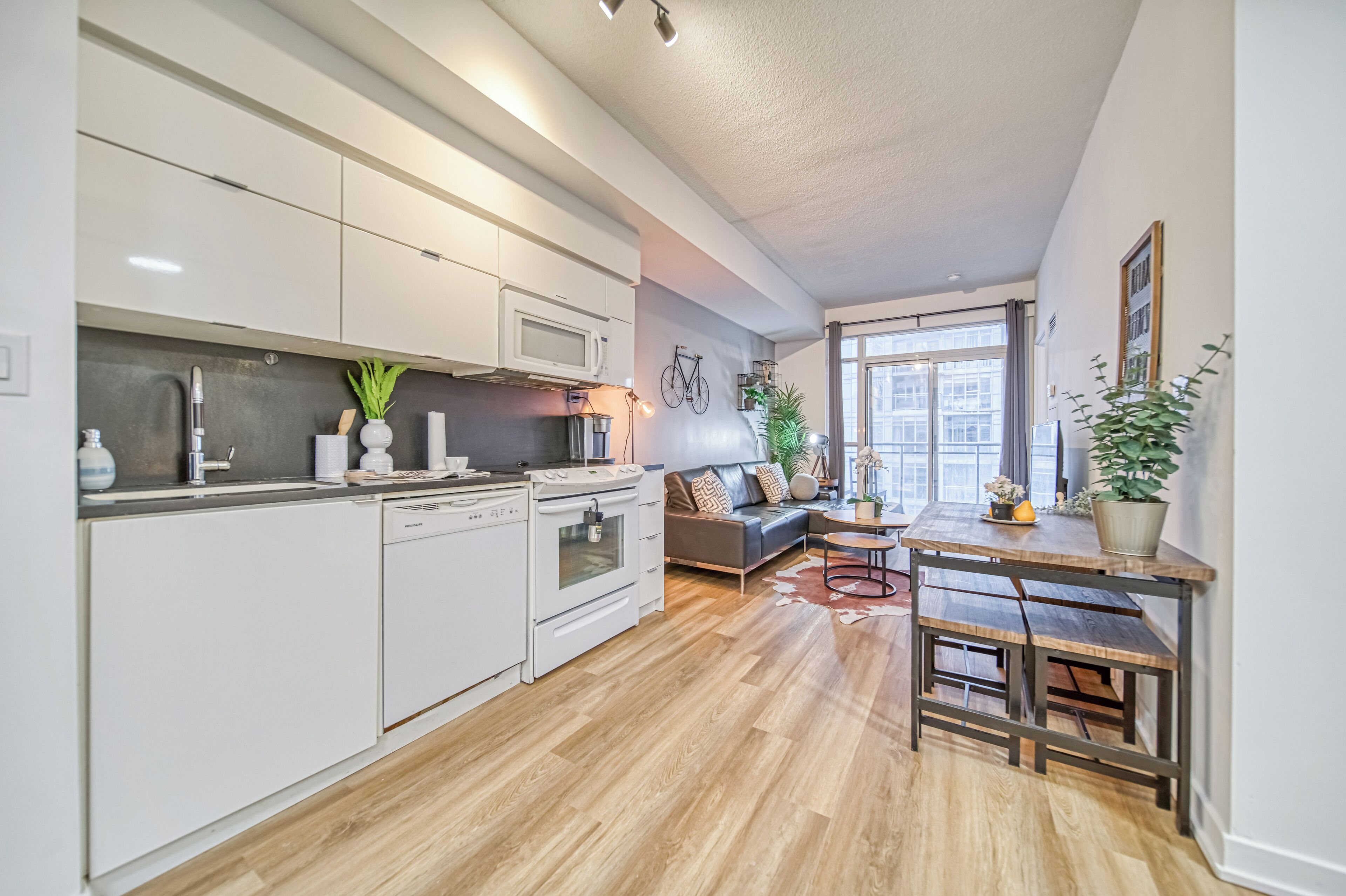 A modern kitchen with white cabinets, a built-in oven, and a microwave. There's a dining area with a wooden table and benches.