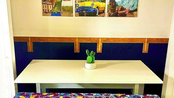 Una habitación con una mesa blanca, un banco con un cojín floral de colores, y una pared con tres cuadros que muestran paisajes naturales.