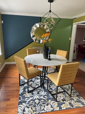 A dining area with a round table, four chairs, a vase with flowers, and a geometric mirror on the wall. - Amazing 4 Bedroom Home Near Airport (Riverdale)