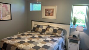 A bedroom with a checkered bedspread, a ceiling fan, a map on the wall, a window with a plant, and a bedside table with a lamp.