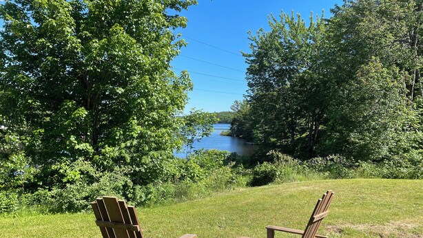 Two wooden Adirondack chairs facing a clear blue lake, surrounded by lush greenery.
