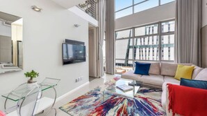 A modern living room with a large window, a sofa, a coffee table, and a glass table with a plant. - Hydro Park Penthouse Loft (Sandton)