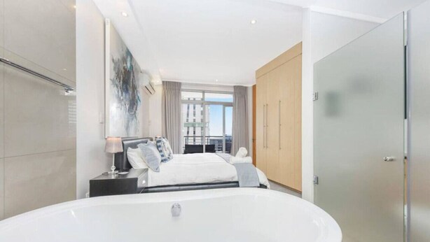 A modern bedroom with a large bathtub, a bed with pillows, a nightstand, and a window with curtains. - Hydro Park Penthouse Loft (Sandton)