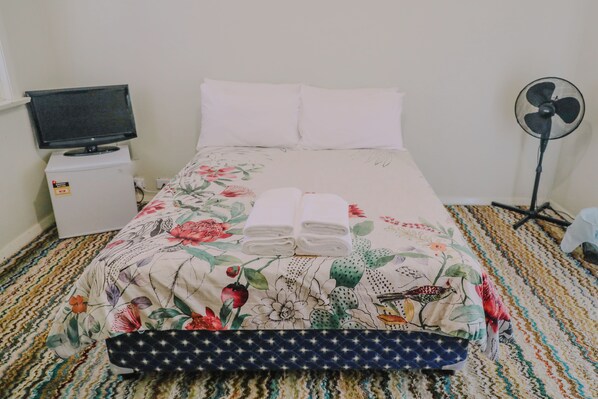 A bed with floral bedding, two white pillows, and two folded towels. A television is mounted on a small white cabinet to the left. A standing fan is positioned to the right of the bed. - Triple Room With Shared Bathroom (Capertee)