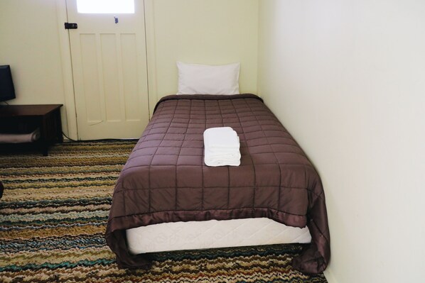 A neatly made bed with a brown quilt and white pillows, a television on a wooden stand, and a patterned carpet.