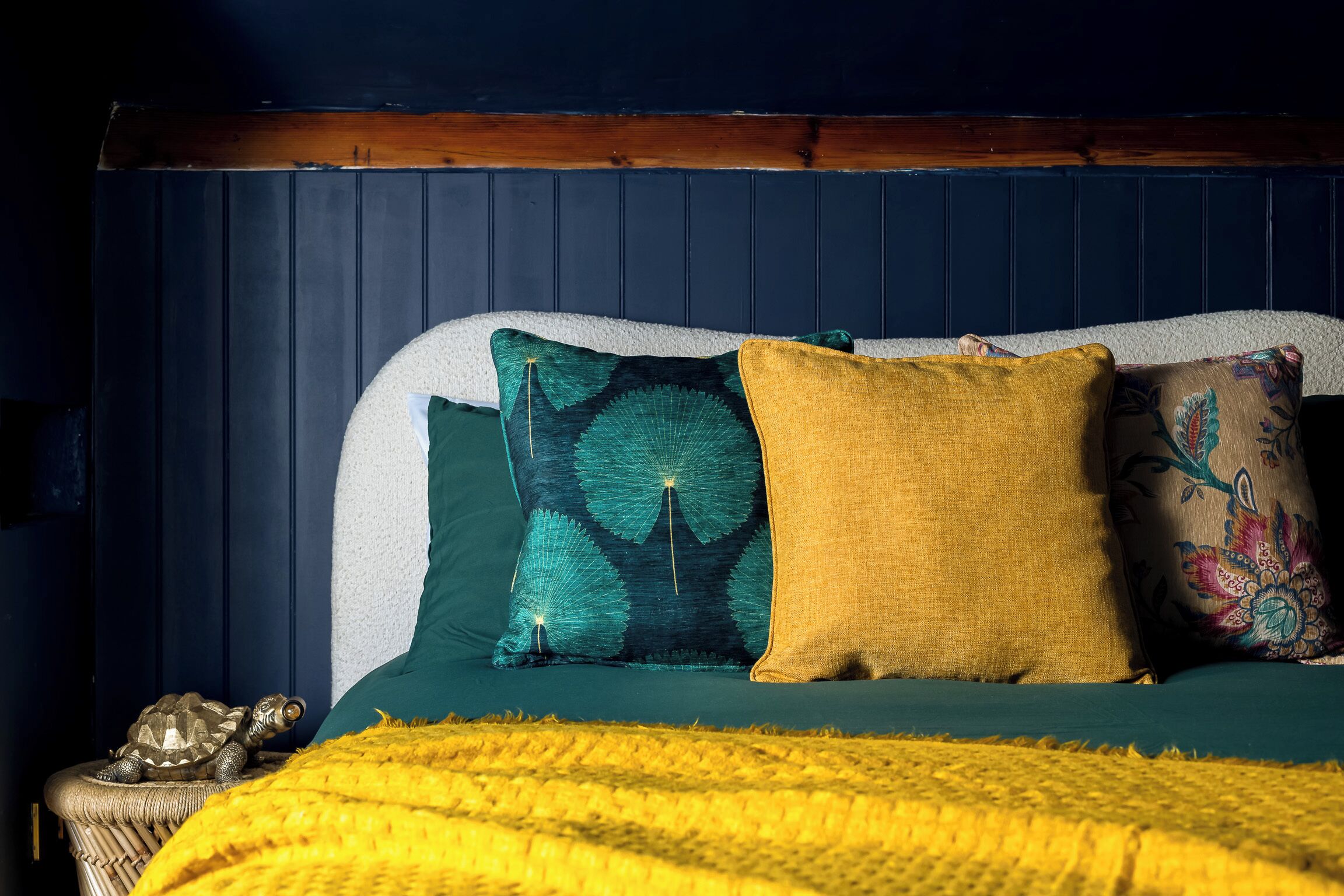 Een bed met een groene sprei, een gele deken en drie kussens met verschillende patronen.