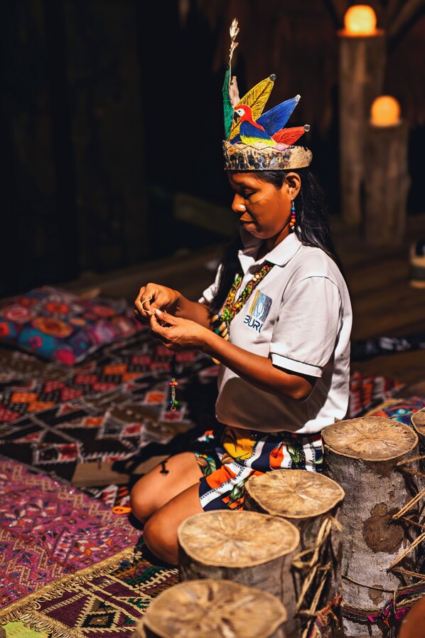 Une personne assise sur un tapis à motifs, entourée de tabourets en bois et d’une couverture tissée aux couleurs vives.