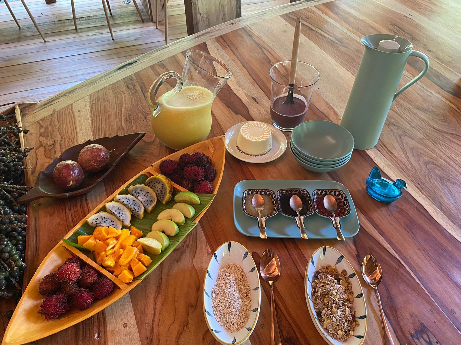 Una mesa de madera con diversos alimentos y bebidas, incluyendo frutas, una jarra de jugo y tazones de cereal.