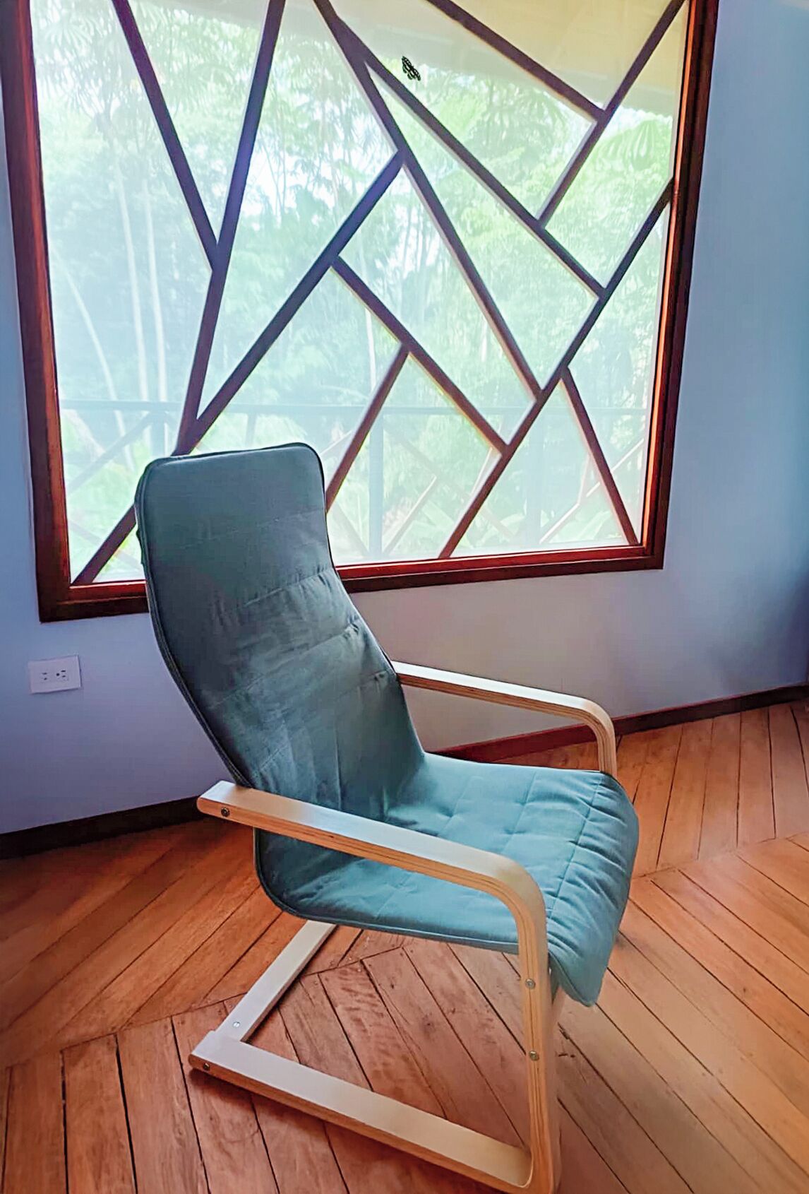 Ein smaragdgrüner Sitzhocker mit Holzgestell in einem Raum mit Holzboden und einem großen Fenster mit geometrischem Muster.