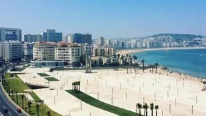 A coastal cityscape with a wide sandy beach, modern buildings, and a clear blue sky. - Siréna Del Tánger (Tanger)